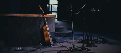 An acoustic guitar rests beside microphone stands on a dim stage.