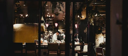 Chefs prepare food inside a restaurant kitchen viewed through large windows.
