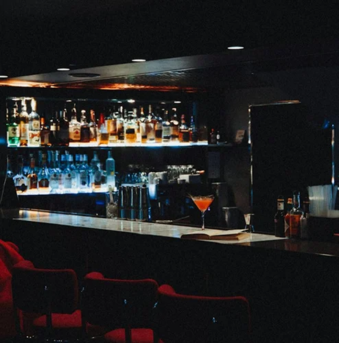A dark cocktail bar features illuminated liquor bottles, red seating, and a drink on the counter.